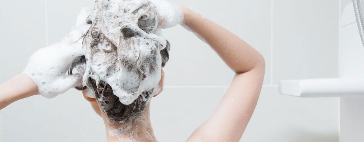 Imagem de uma mulher a tomar banho com a cabeça cheia de espuma de champô.