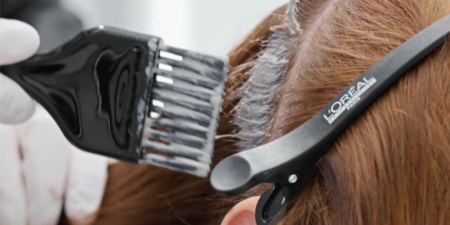 O foco está numa mulher no salão de beleza que tem o cabelo pintado por um profissional, ilustrando como conseguir a cor castanha escura, que está na moda.