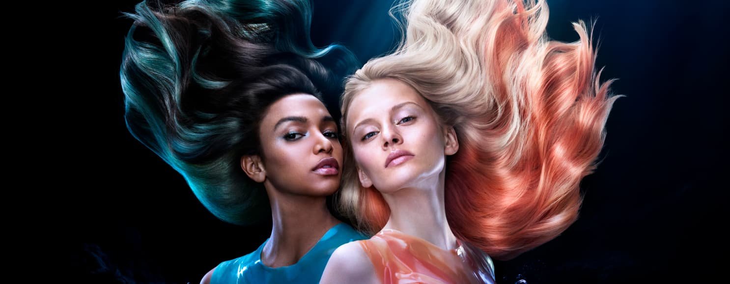 Duas mulheres em pose, uma com cabelo loiro e madeixas laranja, a outra com cabelo escuro e madeixas azuis vibrantes.