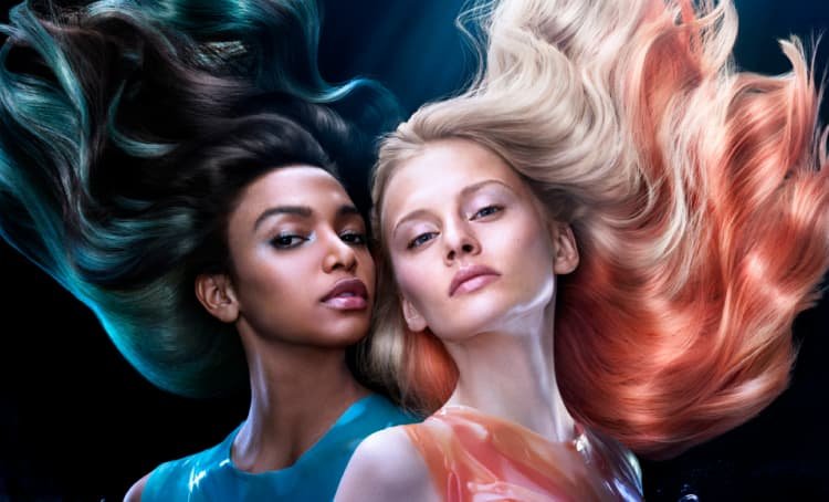 Duas mulheres em pose, uma com cabelo loiro e madeixas laranja, a outra com cabelo escuro e madeixas azuis vibrantes.