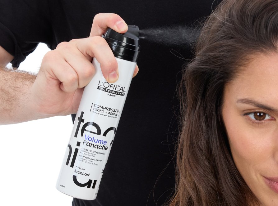 Repare no volume que os cabelos longos de uma mulher ganham enquanto uma cabeleireira profissional utiliza um spray volumizador, seguindo a última tendência de penteados.